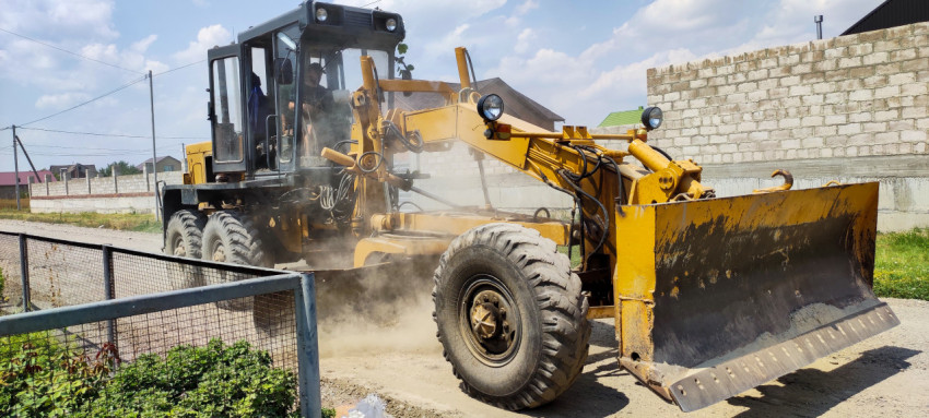 Автогрейдер с отвалом поднимает пыль на просёлочной улице