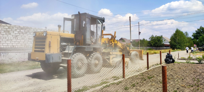 Автогрейдер у сетчатого забора на пыльной дороге в частном секторе
