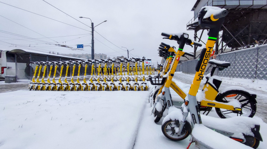 Ряды заснеженных электросамокатов Whoosh на фоне промышленного объекта