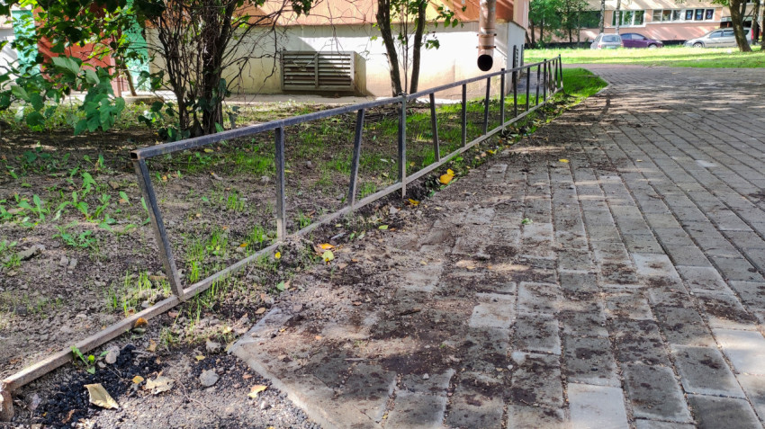 У стены дома рядом с решёткой продуха и водостоком стоит заборчик, а под ним грязь и листья