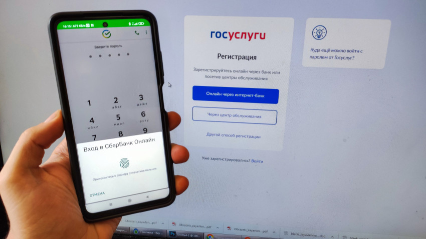 Смартфон с приложением Сбербанка на фоне монитора с открытой страницей регистрации на сайте Госуслуг