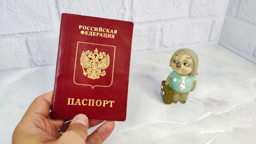 Загранпаспорт РФ в руке и фигурка персонажа с чемоданом