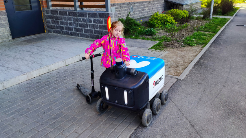 Девочка с самокатом рядом с роботом-доставщиком Яндекс