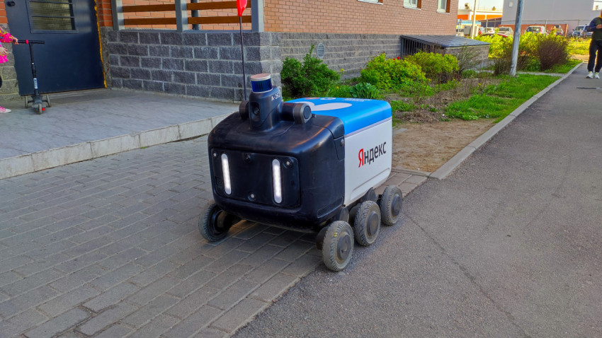 Робот-доставщик Яндекс движется по тротуару у жилого здания