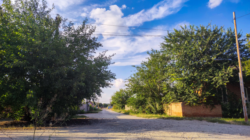Грунтовая дорога в частном секторе с деревьями, кирпичным забором и проводами