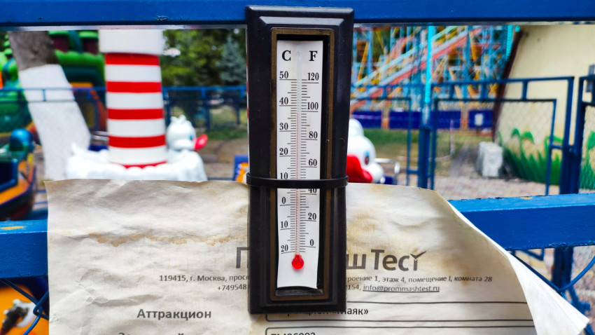 Термометр на заборе на фоне аттракционов в Нальчике