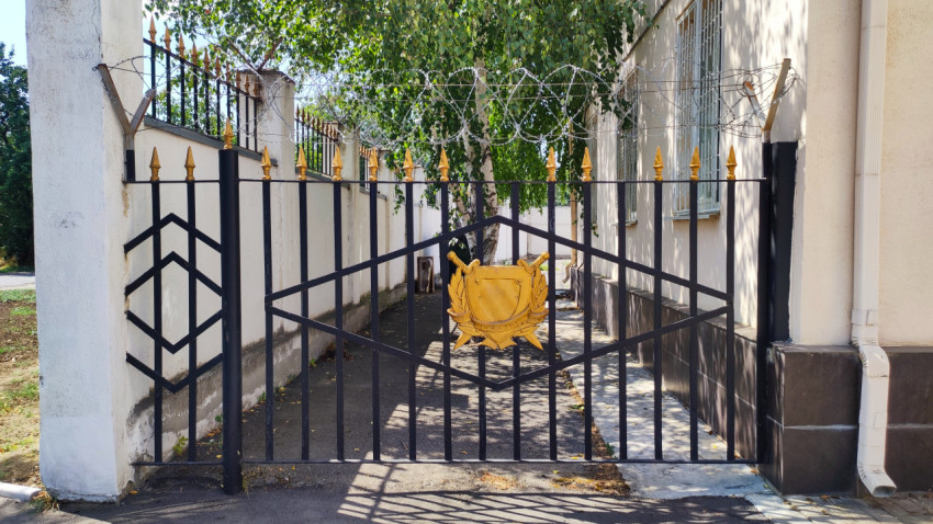 Закрытые ворота с эмблемой на щите и колючей проволокой над забором