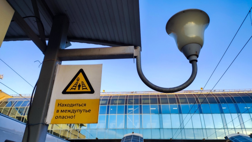 Знак «Находиться в междупутье опасно!» у фонаря на фоне стеклянного фасада вокзала
