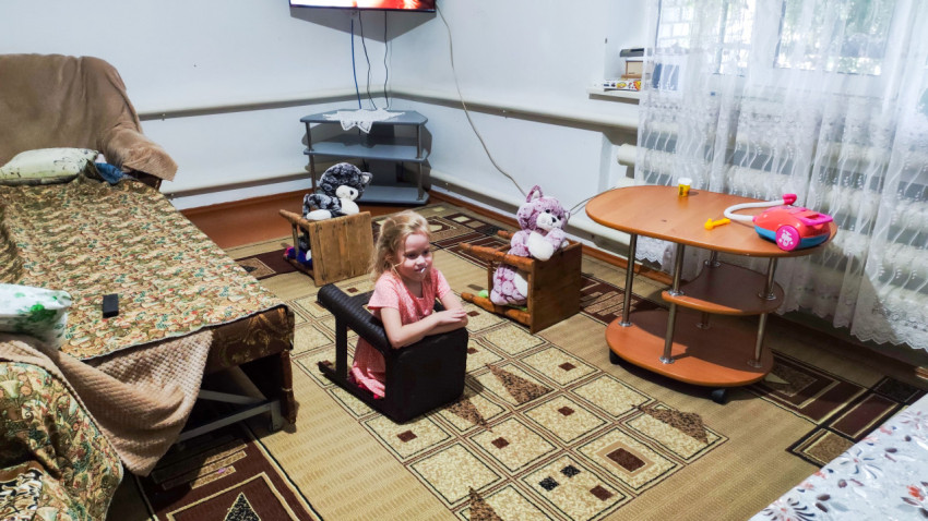 Девочка играет в школу в домашней комнате, сидя за импровизированной партой среди игрушек