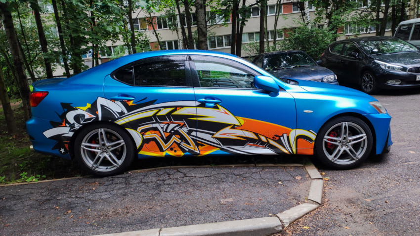 Синий Lexus с графическим винилом на боковинах, стоящий на дворовой парковке рядом с деревьями
