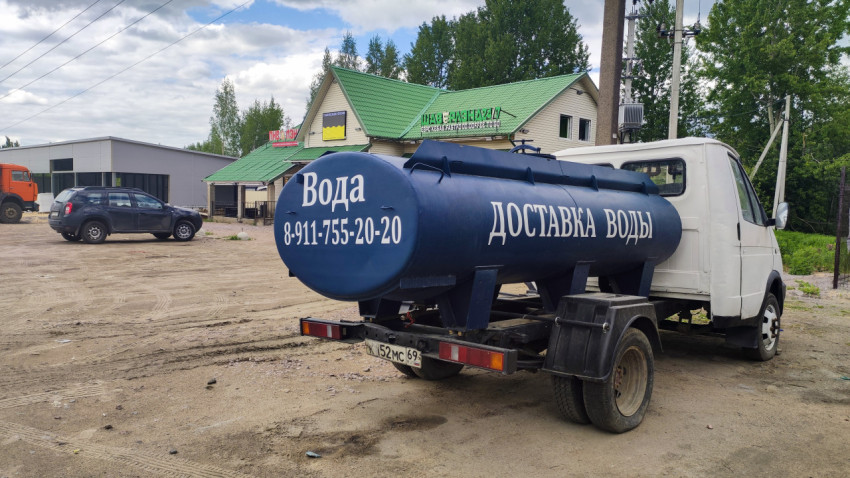 Водовоз с контактным номером компании на фоне придорожных построек