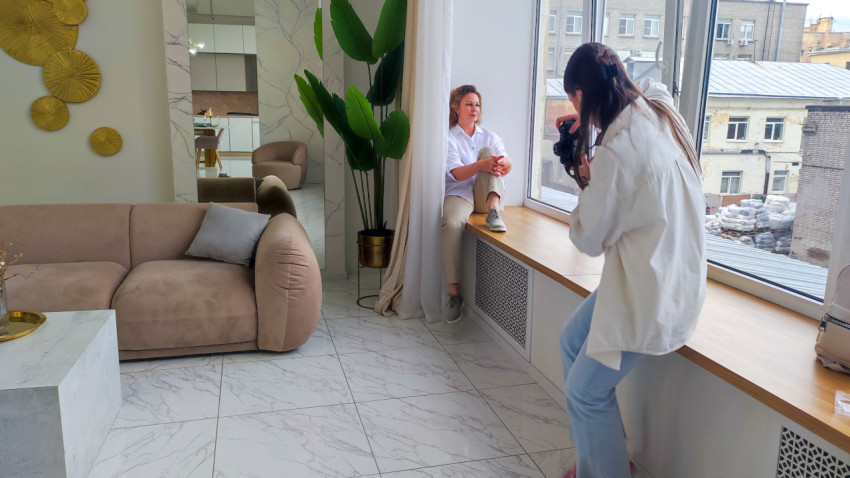 Фотограф снимает женщину у большого окна в жилом интерьере