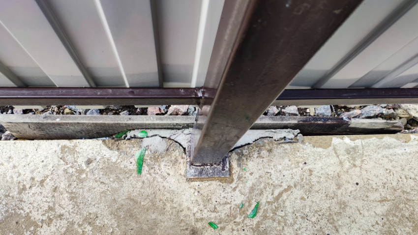 Опора металлического забора с трещинами в бетонной основе