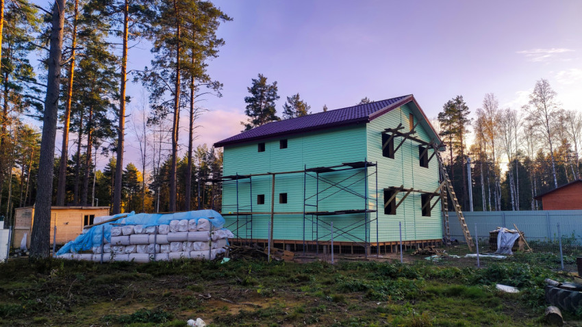 Строящийся дом с зелёным сайдингом и строительными лесами на фоне соснового леса