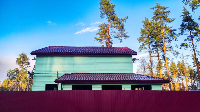 Двухэтажный дом с зелёным фасадом и коричневой металлочерепицей за забором