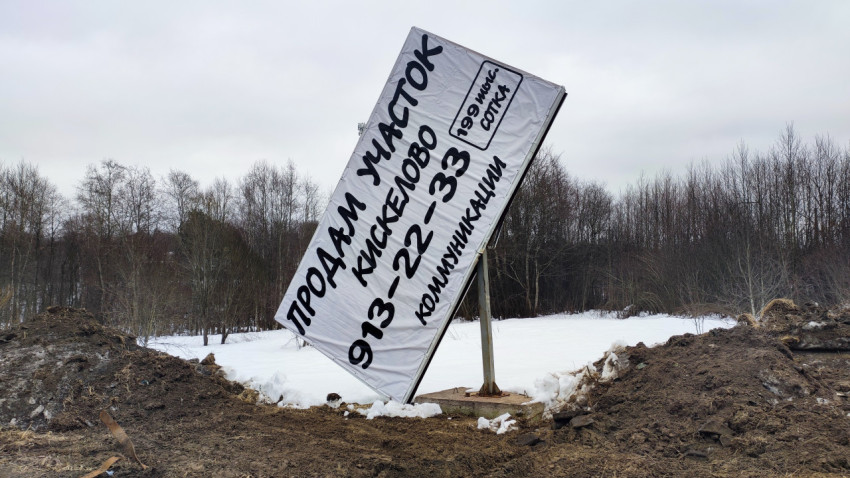 Наклонённый рекламный щит о продаже участка на фоне снега и леса