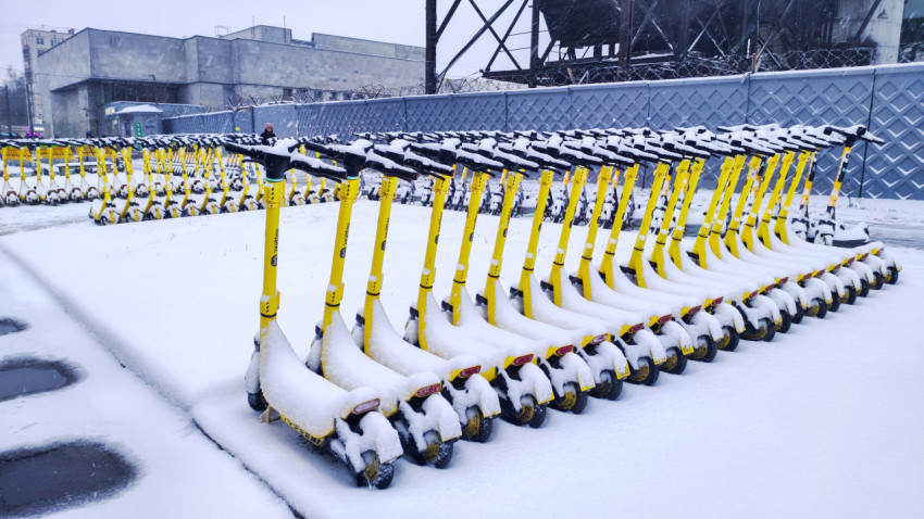 Жёлтые электросамокаты в снегу на открытой площадке перед промышленными сооружениями