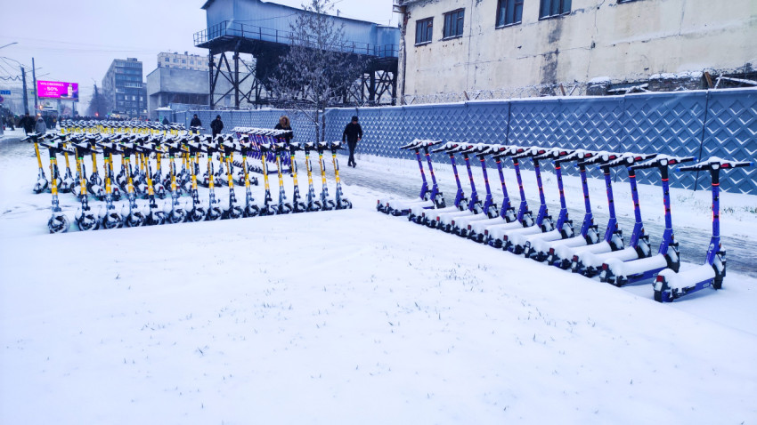 Ряды электросамокатов под снегом вдоль забора промышленной территории