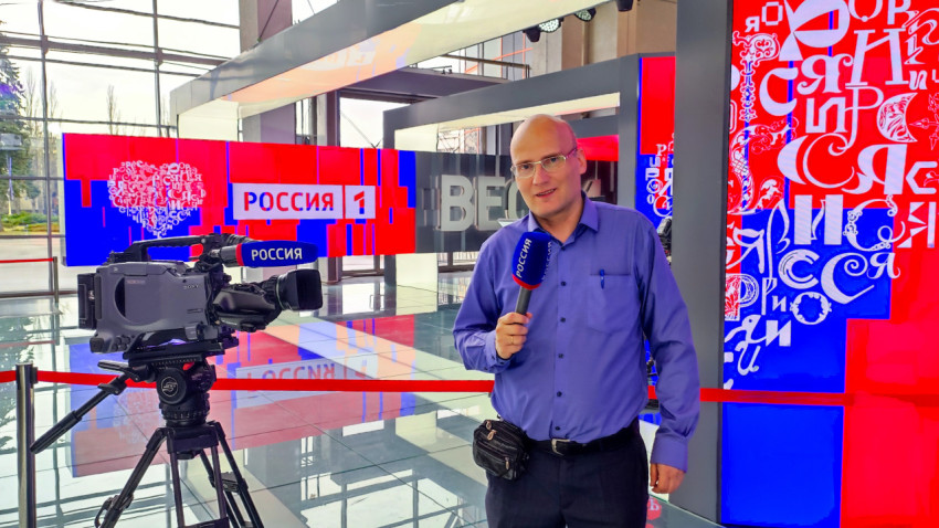 Репортёр с микрофоном «Россия» на фоне студийной зоны «Вести»