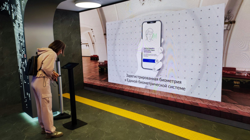 Посетитель взаимодействует с интерактивным терминалом перед экраном о биометрии