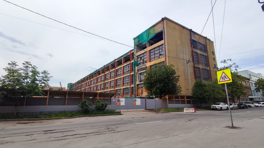 Промышленный корпус с большими окнами за строительным забором и предупреждающим знаком «Дети»