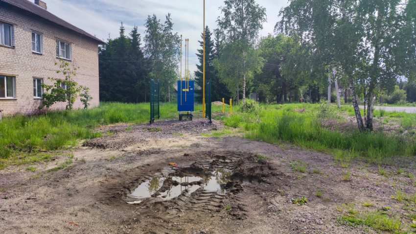 Грязевой проезд с лужей перед ограждённым газовым шкафом и жёлтыми трубами