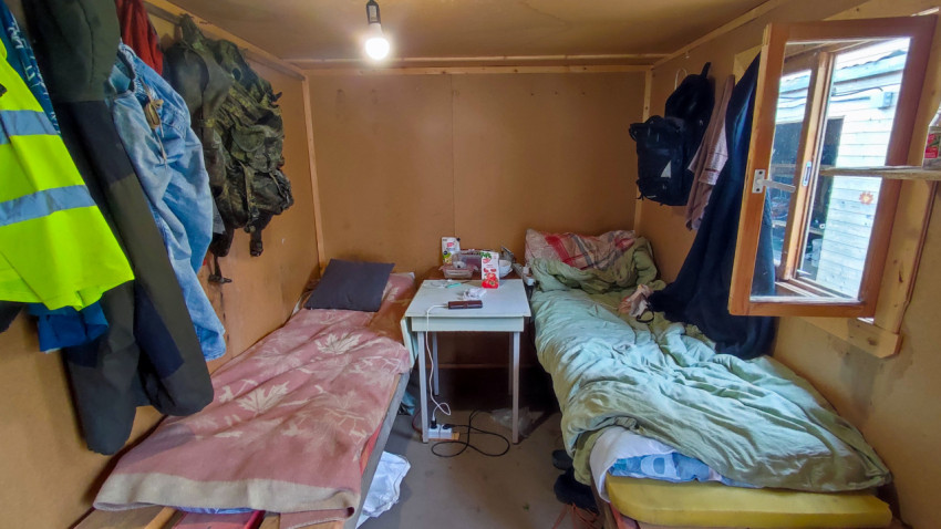 Интерьер бытовки с двумя кроватями, столом и рабочей одеждой на стене