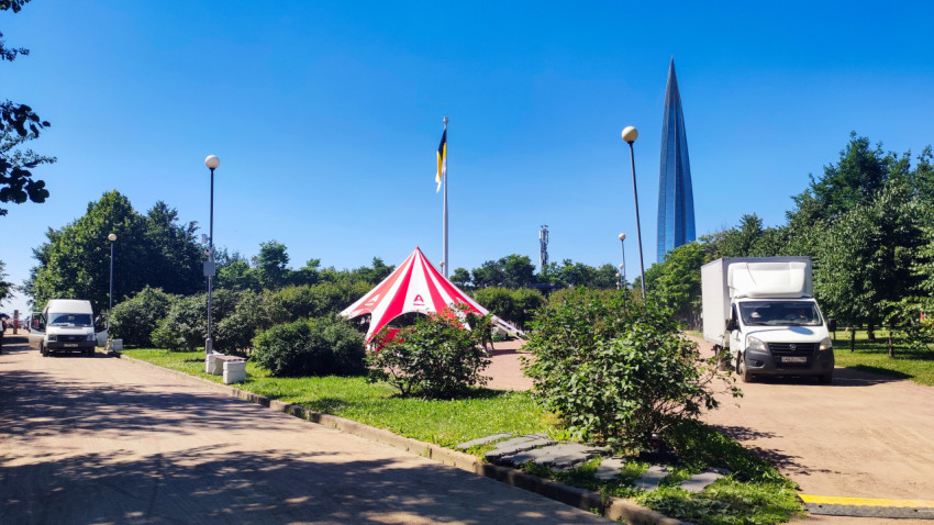 Красно-белый шатёр Альфа-Банк и грузовые фургоны на парковом проезде у кустарников