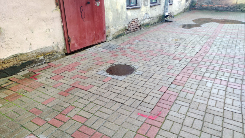 Канализационный люк на дворовой плитке у красной металлической двери и водосточной трубы