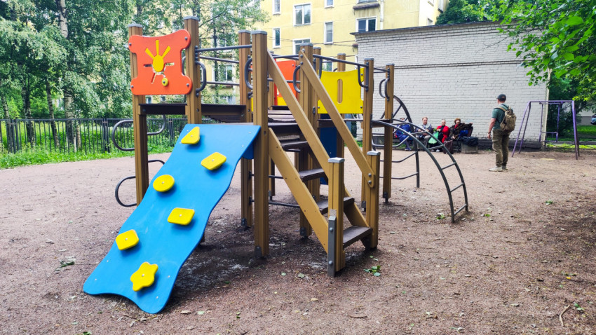 Детский игровой городок со скалодромом во дворе жилых домов и людьми на заднем плане