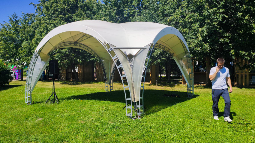 Белый шатер-павильон на газоне и мужчина с телефоном в парковой зоне