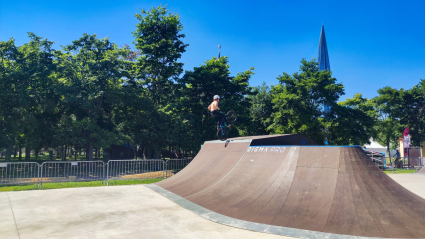 BMX-райдер выполняет трюк на фигуре скейтпарка на фоне деревьев и небоскрёба