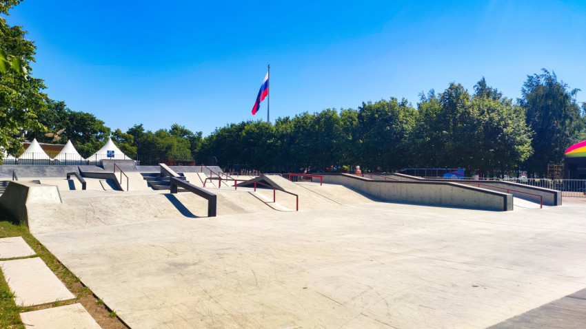 Панорама скейтпарка с рейлами, шатрами и флагштоком на фоне зеленой аллеи