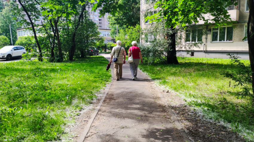 Пожилая пара идёт по тротуару в зелёном дворе многоквартирных домов