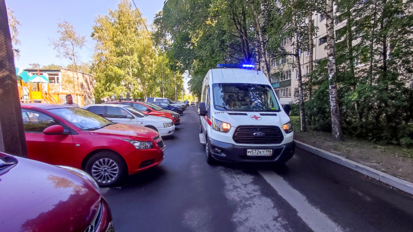 Машина скорой помощи подъезжает по дворовому проезду рядом с парковкой и детской площадкой