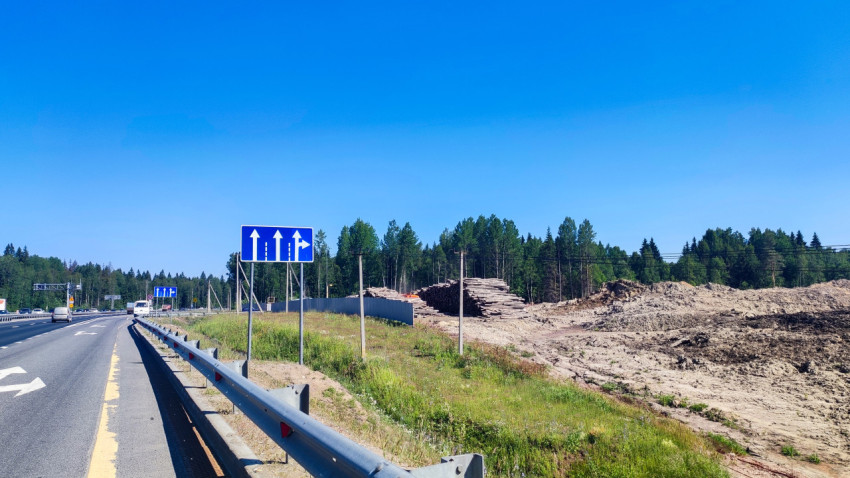 Автомагистраль с отбойником и указателями полос рядом с площадкой складирования брёвен и земляными работами