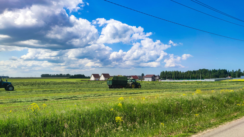 Трактор с прицепом на поле с валками сена и деревней на горизонте