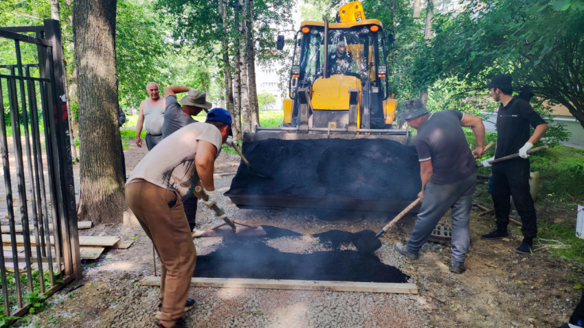 Рабочие распределяют горячий асфальт лопатами перед ковшом экскаватора-погрузчика JCB