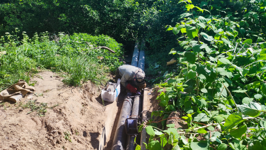 Монтаж коммуникаций в траншее: рабочий у стыка труб и кабелей в густой растительности