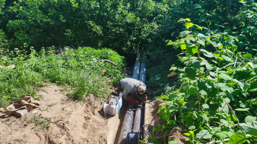 Рабочий выполняет монтаж на трубопроводе в открытой траншее среди зелёных зарослей