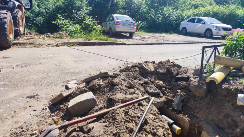 Раскопанный участок дороги с грунтом и инструментом возле колес погрузчика и припаркованных машин