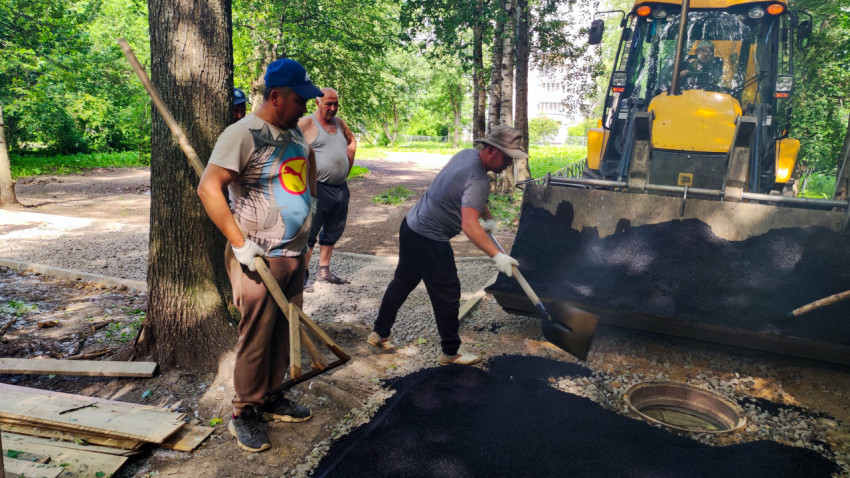 Рабочие с лопатами и граблями разгружают асфальтовую смесь из ковша JCB во дворе у деревьев