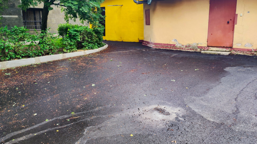 Свежий асфальт у стены жилого дома с жёлтыми воротами и следами ремонта покрытия