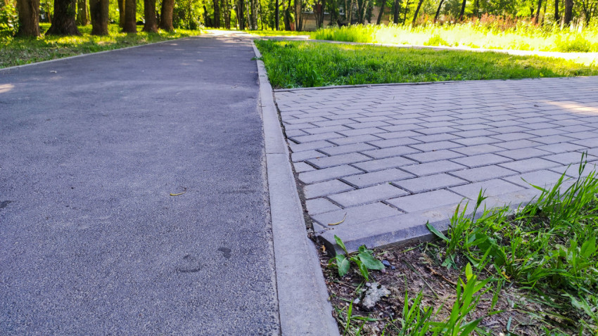 Пешеходная дорожка и плиточная площадка, разделённые бордюром, в зелёной парковой зоне