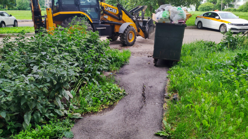 Асфальтовая дворовая дорожка со стрескавшимся покрытием рядом с мусорным контейнером и экскаватором-погрузчиком JCB