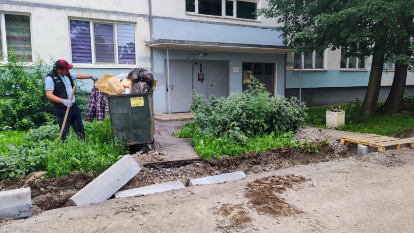 Рабочий с лопатой у переполненного контейнера на фоне подъезда жилого дома и раскопки через проезд