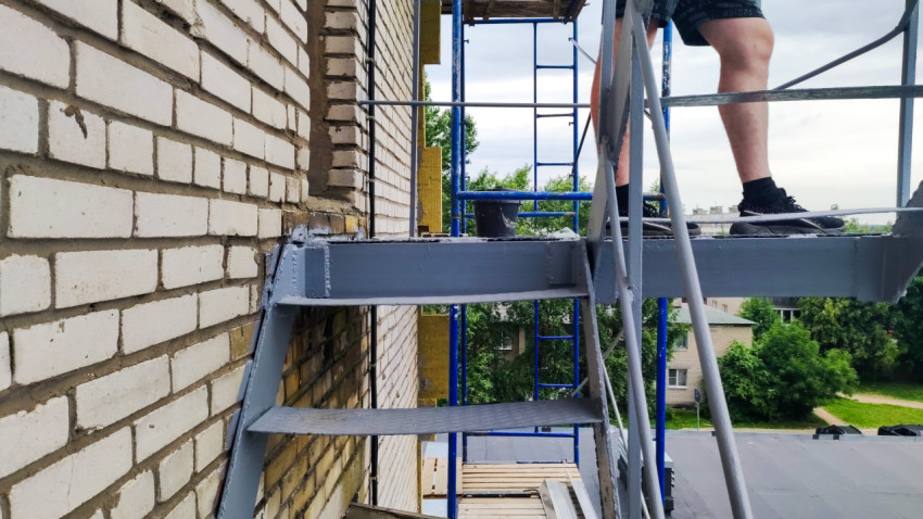Металлическая лестница и площадка у кирпичного фасада рядом со строительными лесами