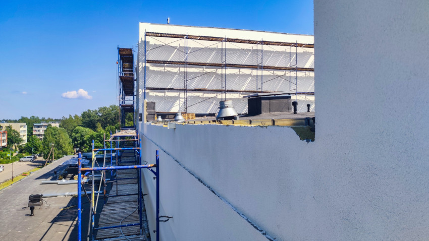 Вид с лесов на белый фасад и край кровли с открытым слоем утеплителя и вентиляционными выходами