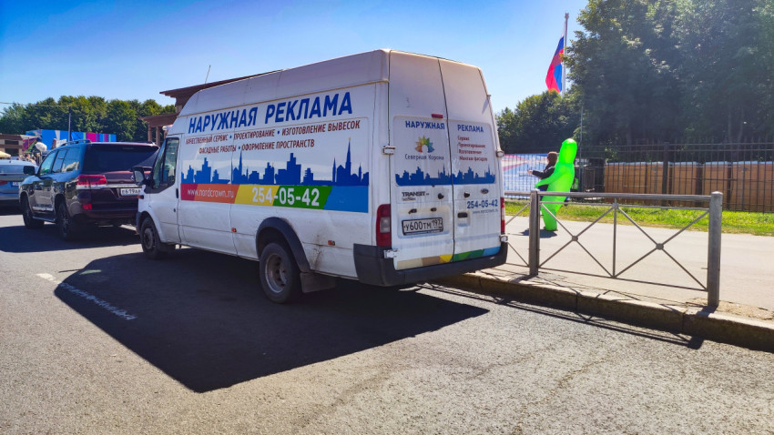 Фургон Ford Transit с надписью «Наружная реклама» припаркован у тротуара в солнечный день