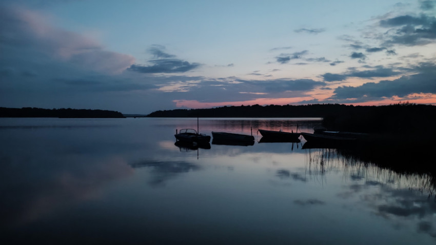 Закат над озером с лодками у берега и отражением облаков в воде
